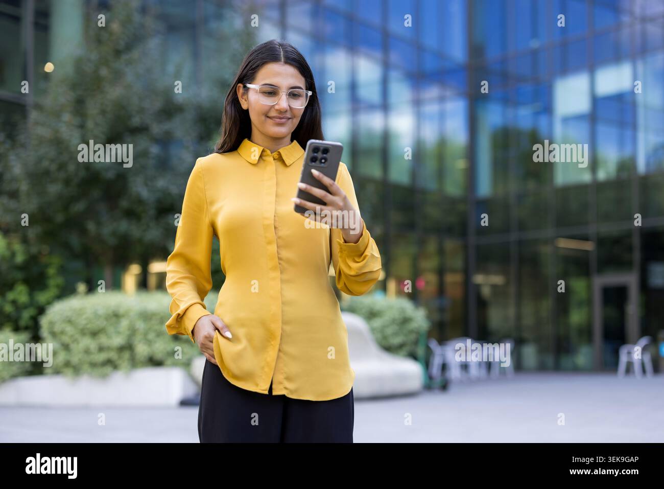 Professional woman in yellow blouse and clear glasses checking her smartphone, standing in front of a modern glass building, utilizing mobile technology for communication Stock Photo