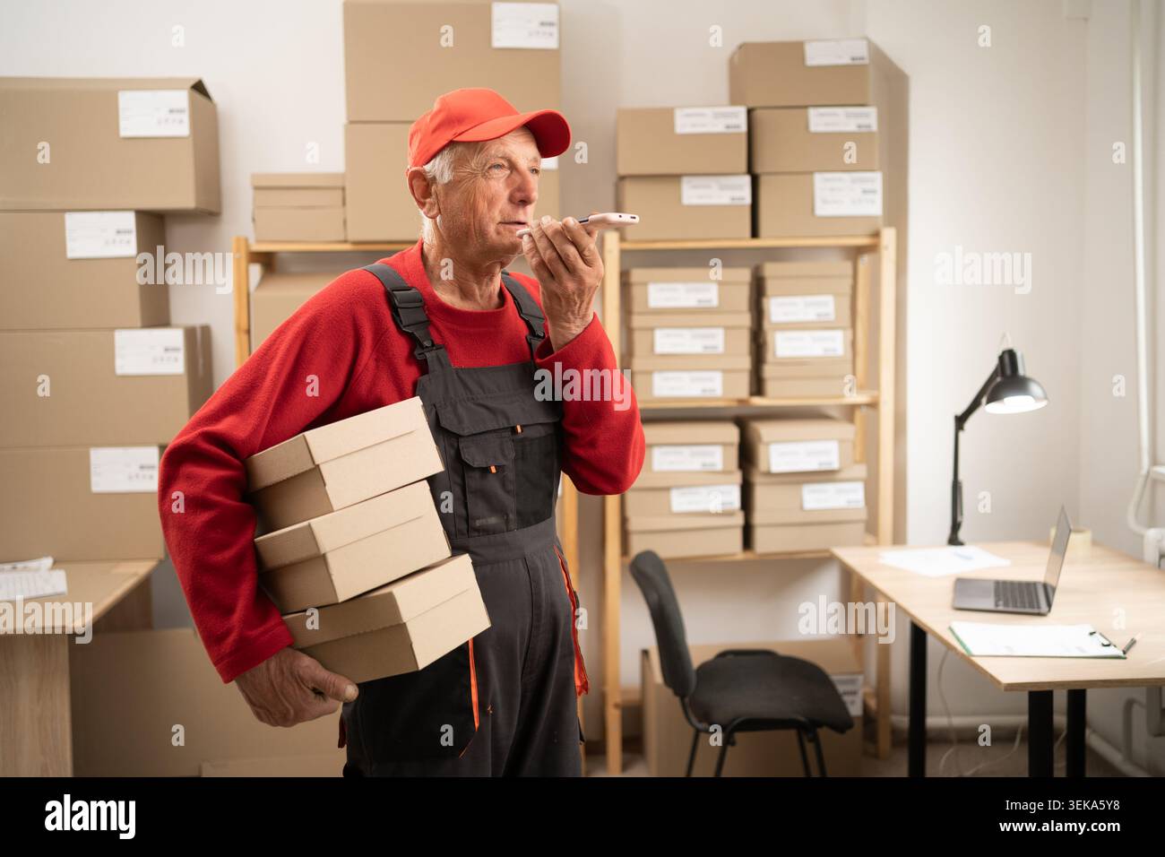 Elderly postal worker holding parcels in warehouse using AI voice assistant Stock Photo