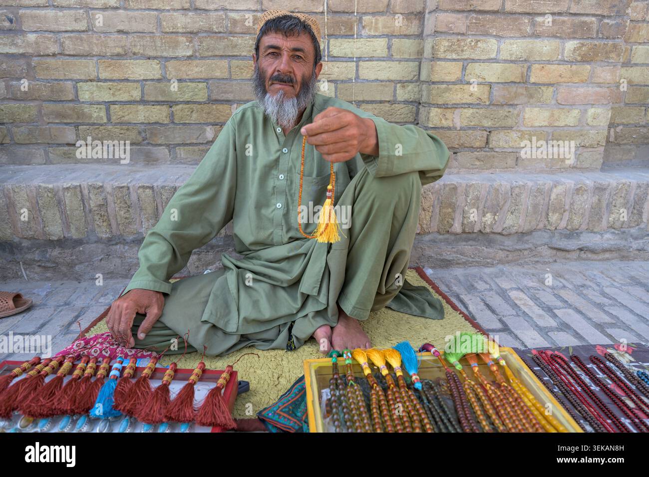 Man selling strings of traditional prayer beads, sitting on a mat in a market setting outside the great mosque of Herat. Stock Photo