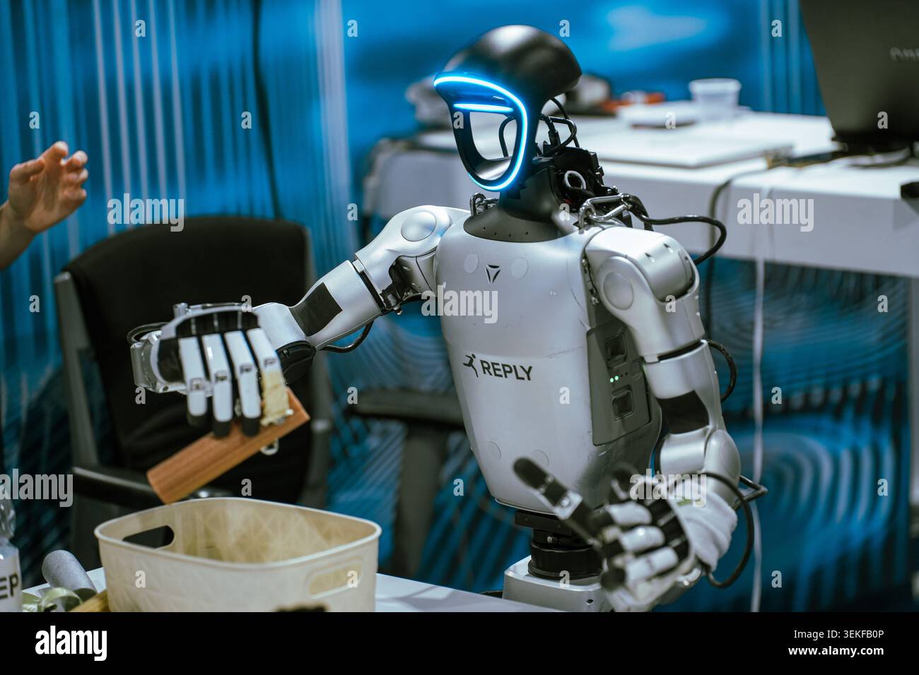 A humanoid robot with a glowing blue visor interfaces with an object over a container during a live automation and programming demonstration at a tech Stock Photo