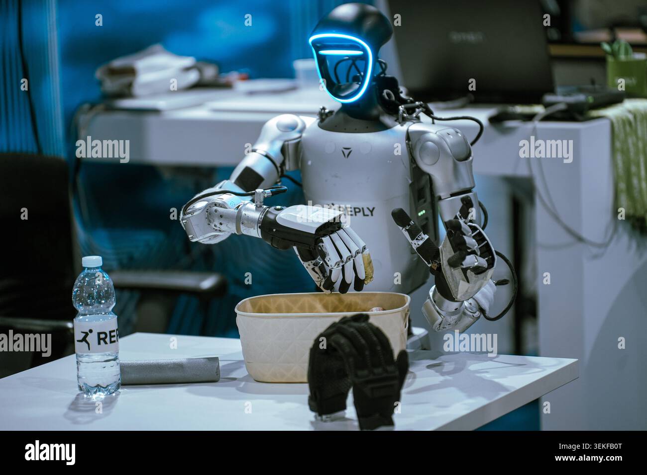 A humanoid robot with a glowing blue visor interfaces with an object over a container during a live automation and programming demonstration at a tech Stock Photo