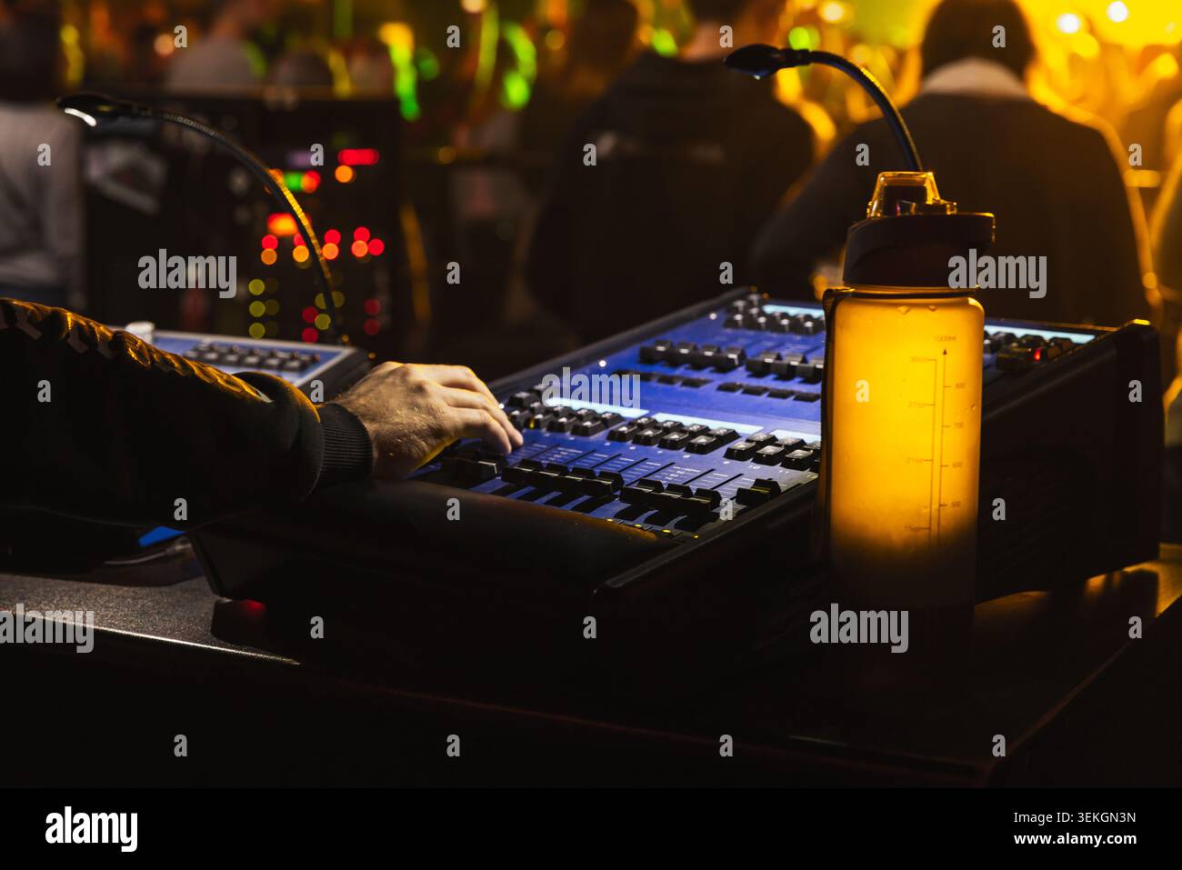 Experienced audio technician operates a mixing console during a live concert, adjusting faders and levels under stage lighting while a glowing water b Stock Photo