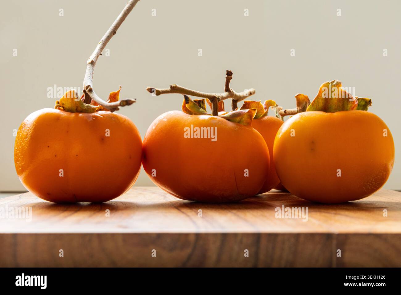 Three ripe persimmons arranged on a wooden cutting board in soft natural light. Seasonal autumn fruit with warm colors and minimalist composition. Stock Photo
