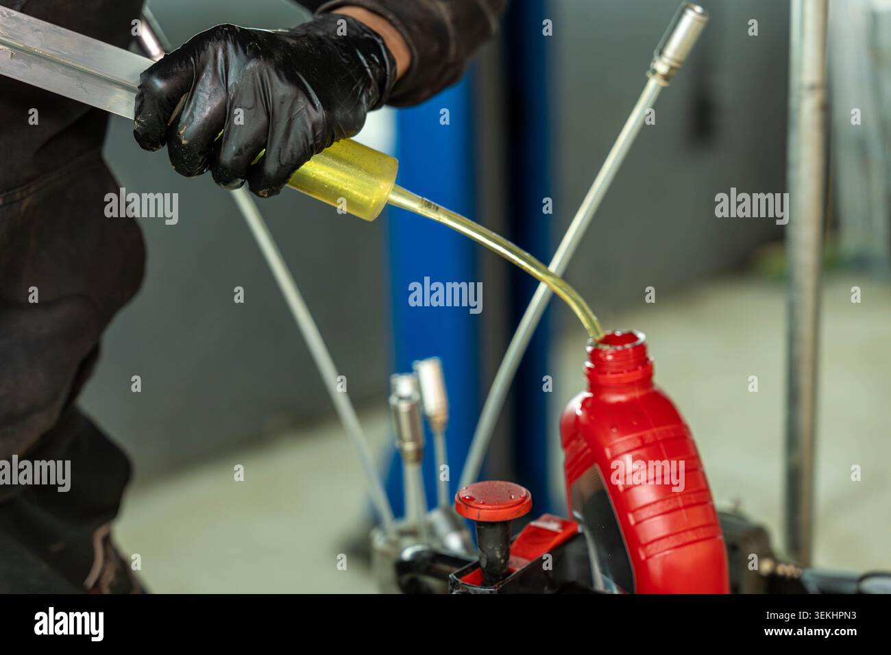 A close up view of a worker wearing black gloves pouring yellow liquid into a red plastic container in an industrial workshop, with blurred tools Stock Photo