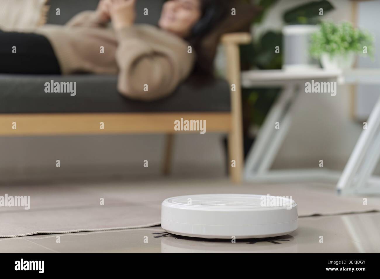 A robot vacuum operating on a tiled floor in a contemporary living room while a person relaxes on a couch in the background Stock Photo