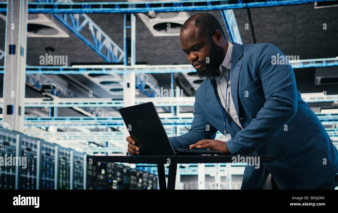 Programmer at server farm workplace desk using laptop, doing network management. Data center IT expert overseeing IT infrastructure supercomputers with software, typing on notebook keyboard, camera A Stock Photo