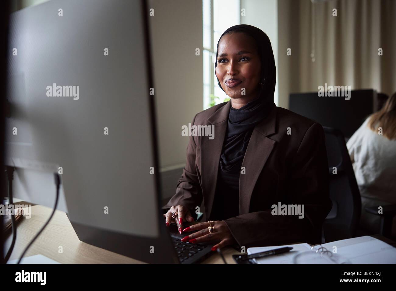 Smiling female tech expert using laptop and computer while sitting at desk in office Stock Photo