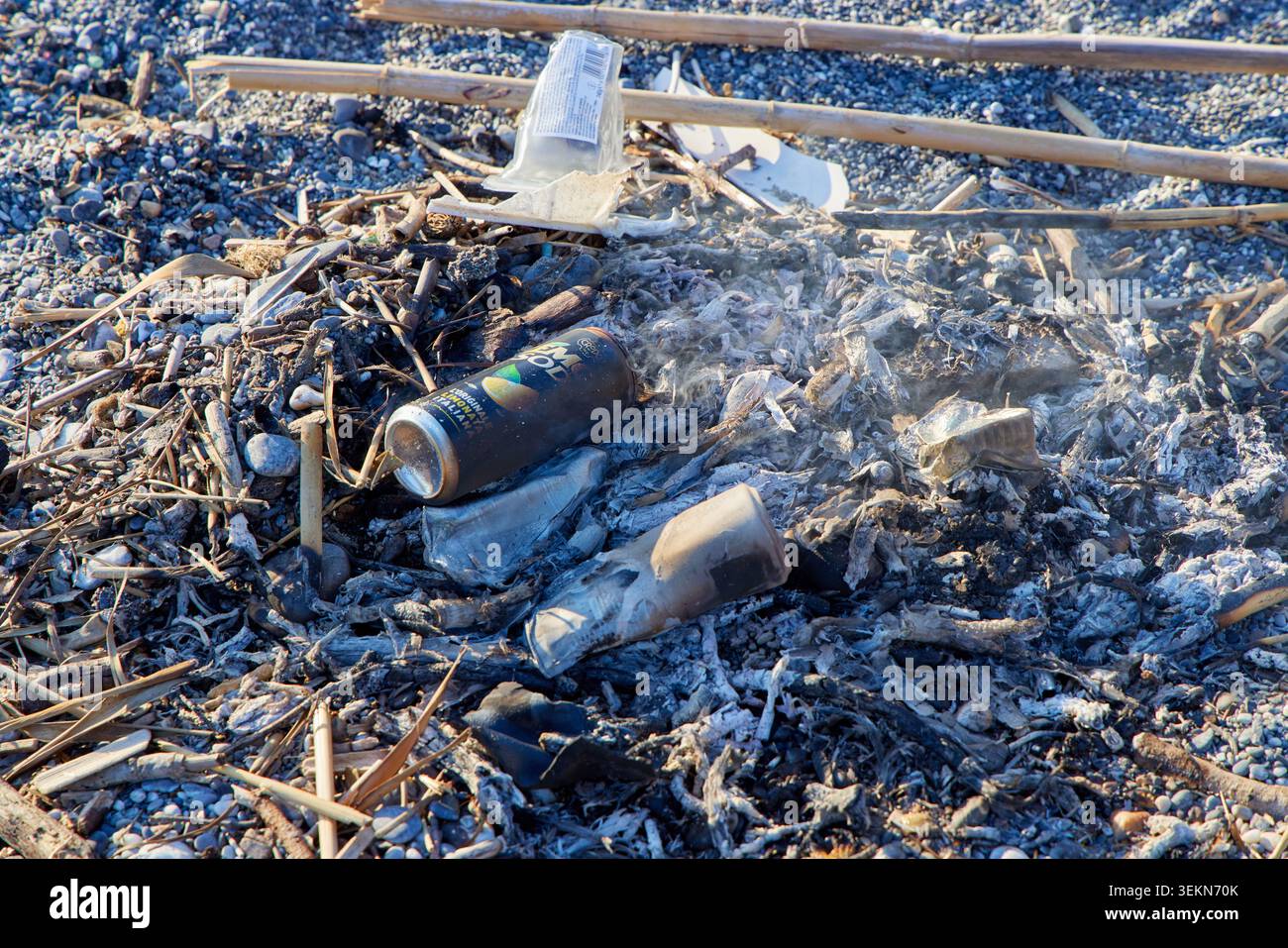 Discarded aluminum cans and plastic trash burnt in a campfire on a pebble beach close up. Stock Photo