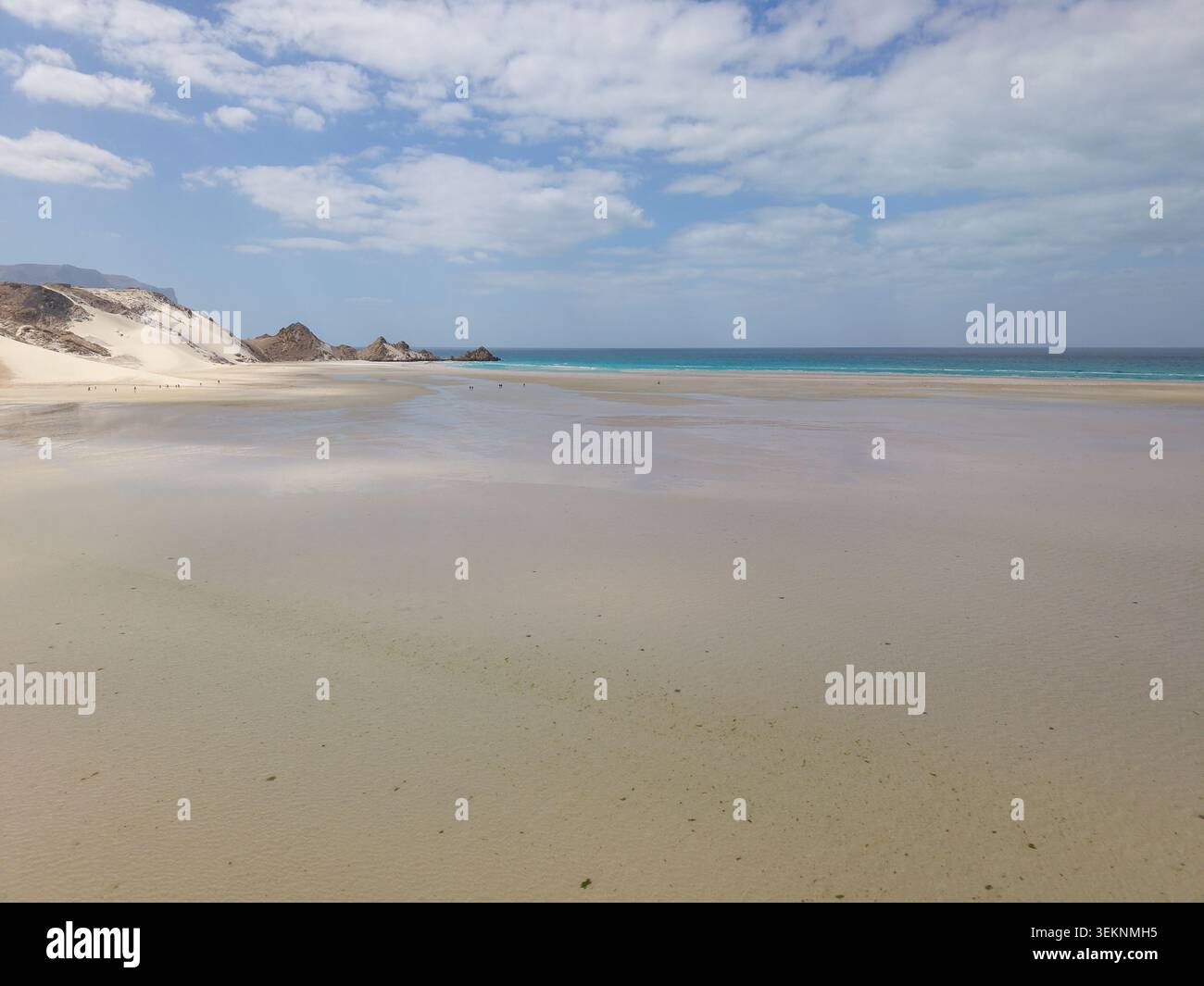 Wide sandy beach with shallow wet shore, turquoise sea, pale dunes and rocky cliffs under a soft cloudy sky. Calm coastal landscape with open space an Stock Photo