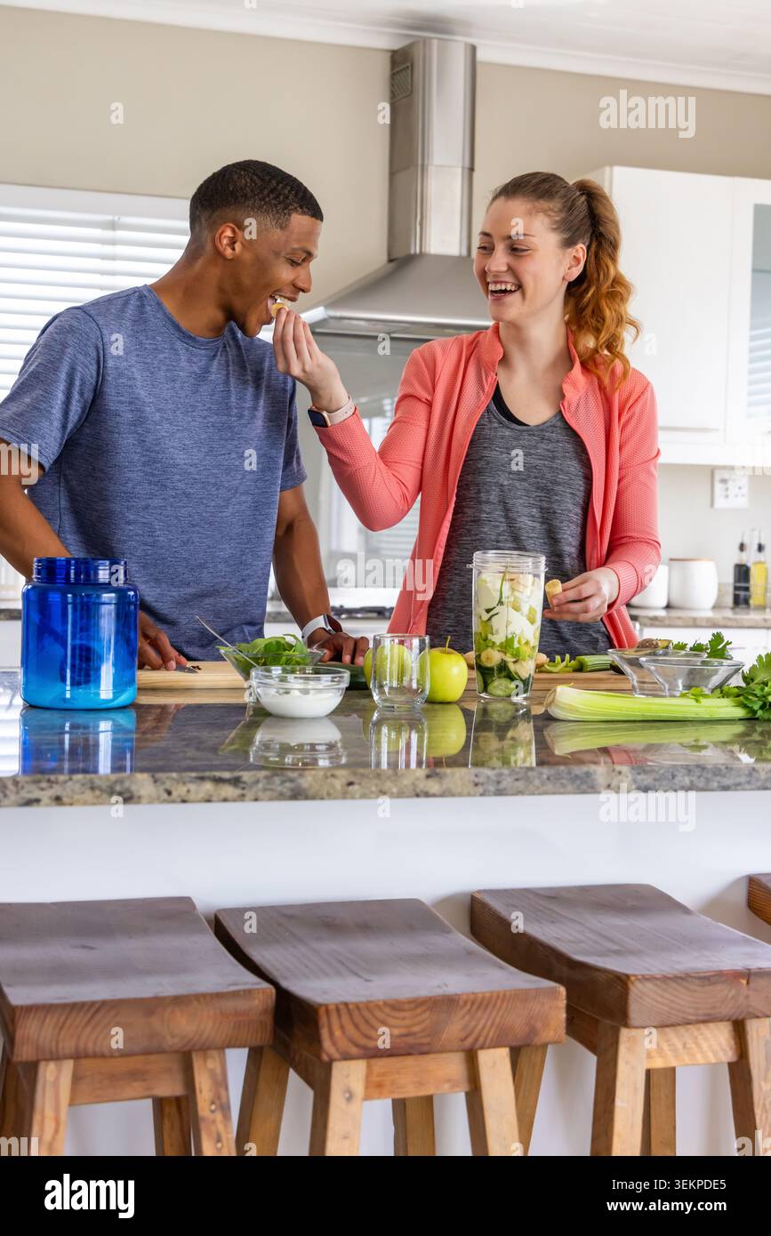 Diverse couple feeding each other while preparing green smoothie in home kitchen with blender Stock Photo