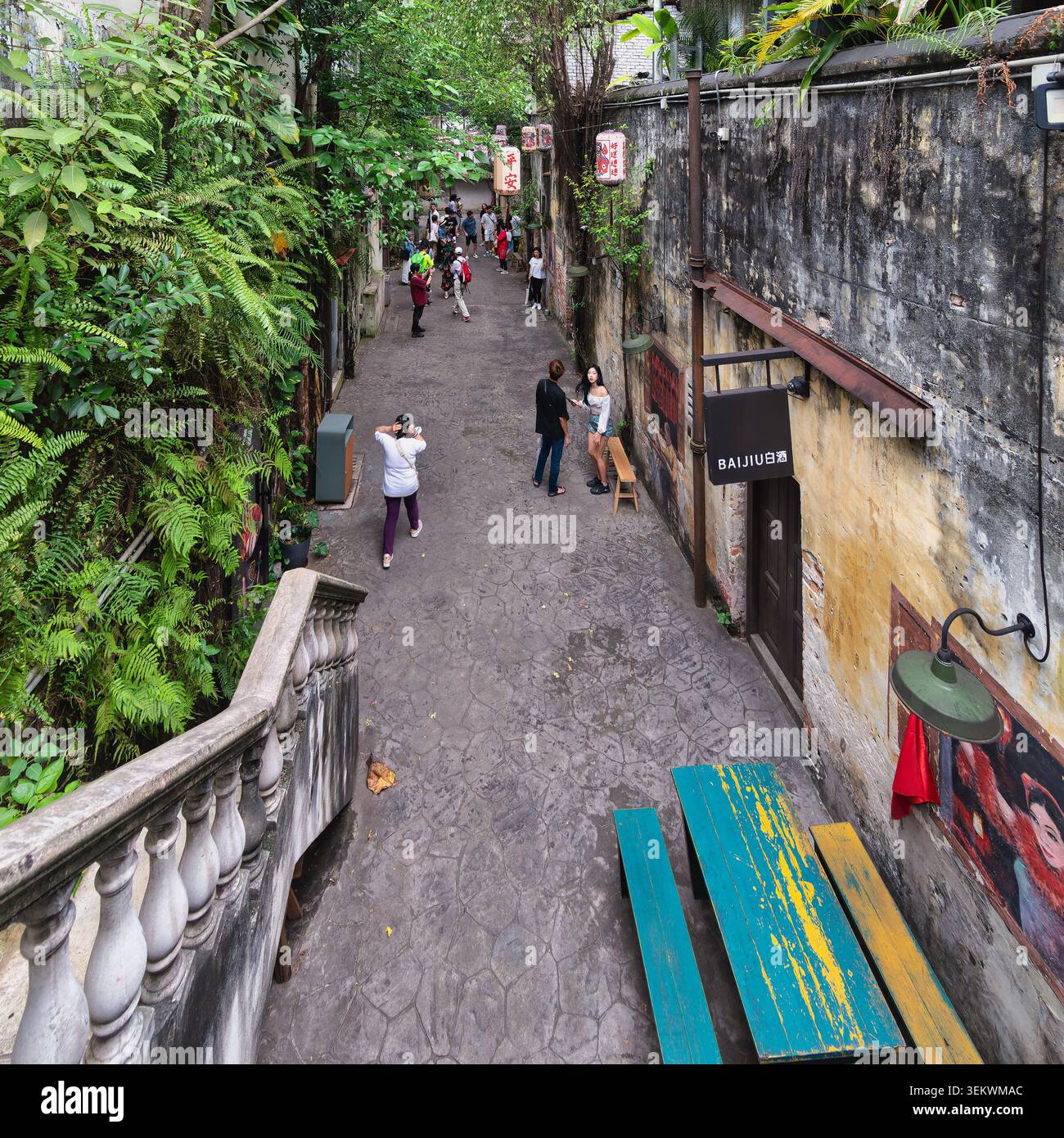 Kuala Lumpur, Malaysia - February 21, 2025: Kwai Chai Hong alley with people, lush ferns, old walls, Chinese lanterns, and colorful benches, showcasing cultural charm Stock Photo
