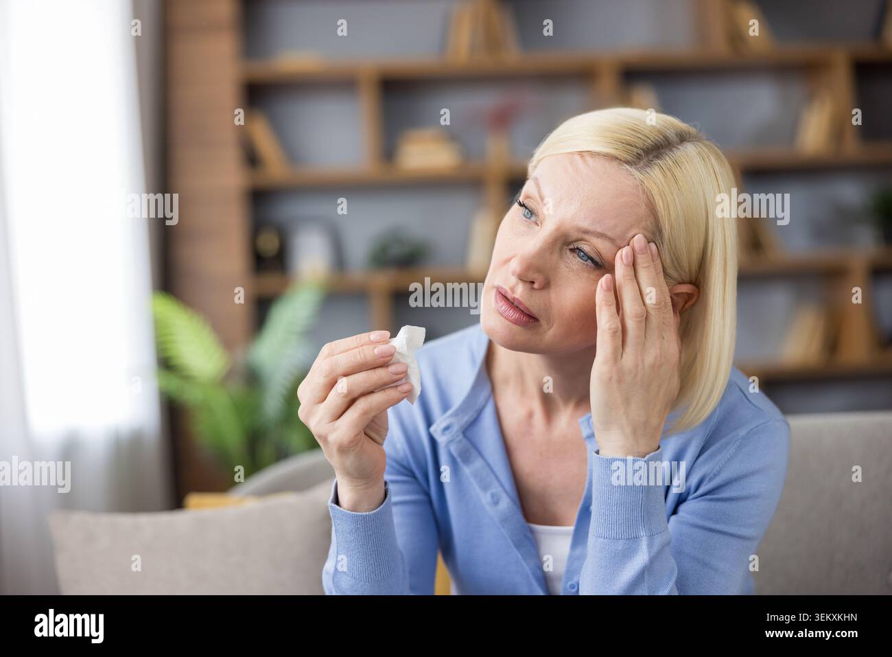 Mature blonde woman suffering from severe migraine and stress, touching her head with a hand, holding a white tissue, looking sad and tired, sitting indoors on a couch Stock Photo