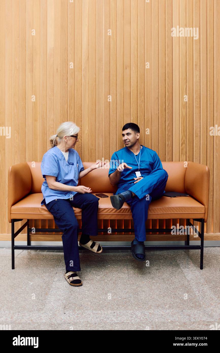 Young male doctor talking with senior female coworker while sitting on sofa in hospital lobby Stock Photo