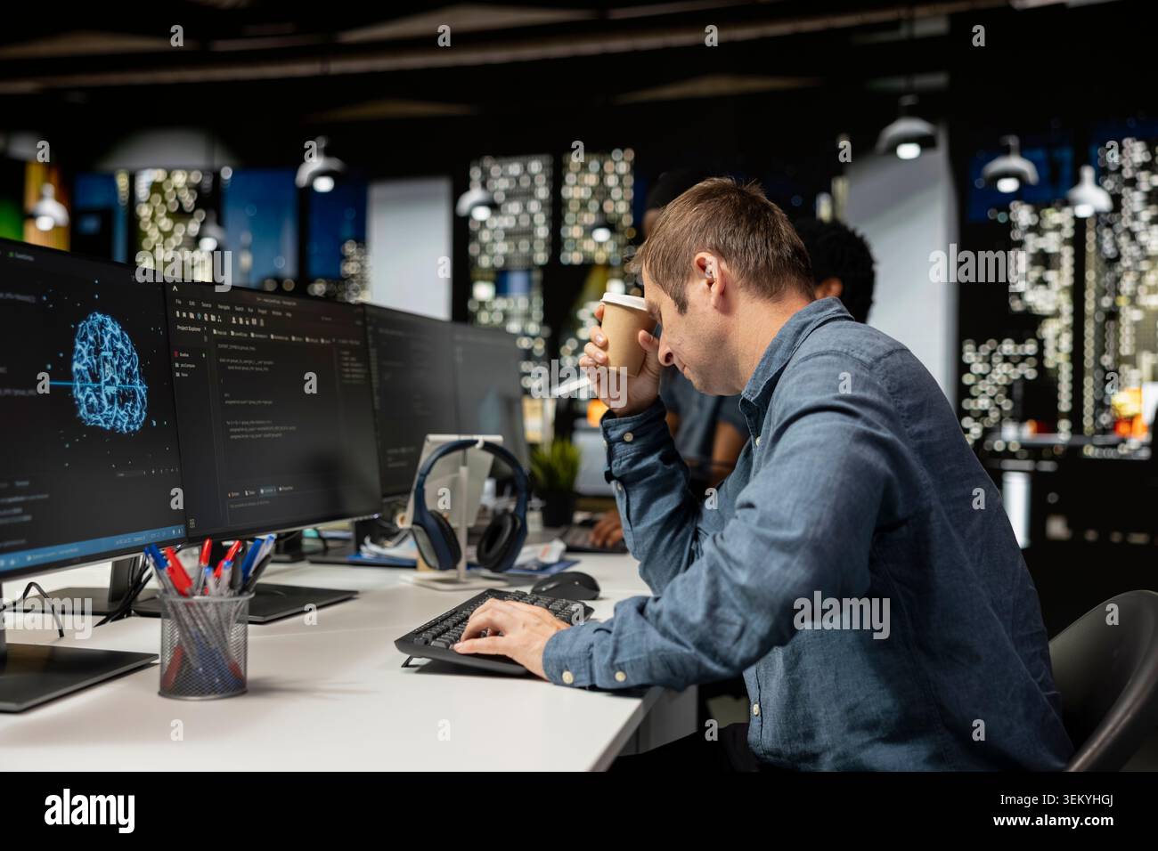 Tired man drinks coffee while utilizing AI on computer to optimize performance. Exhausted worker enjoys caffeinated beverage in tech company office while training deep learning models Stock Photo