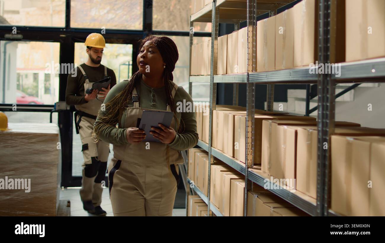 Revealing shot of woman in warehouse inputing updated products info on online shopping website using tablet, walking through retail storage hub aisles checking labels on wares Stock Photo