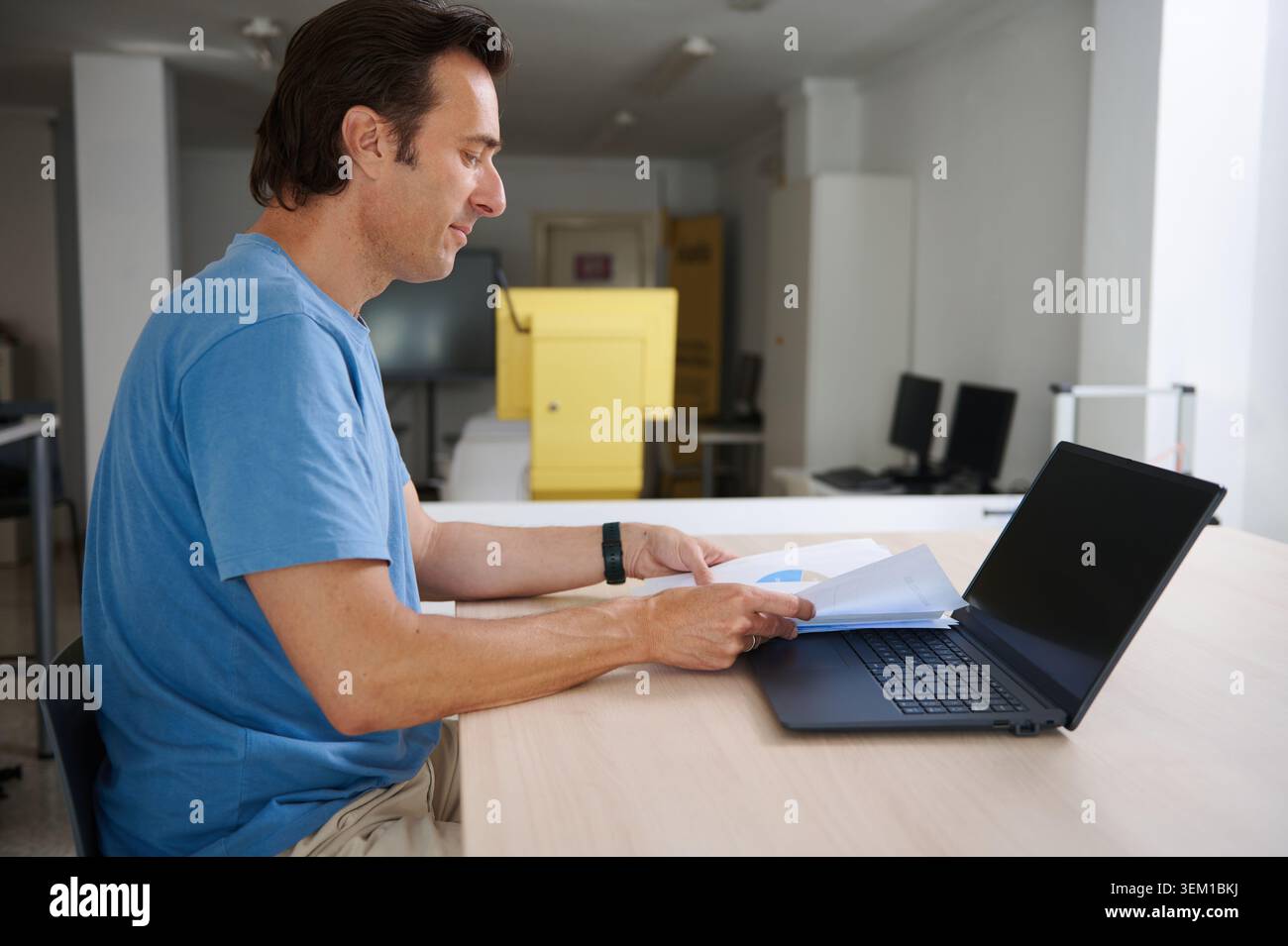 Man seated at office desk, reviewing printed reports and charts while using a laptop - calm focused small-business or remote-work scene with modern wo Stock Photo