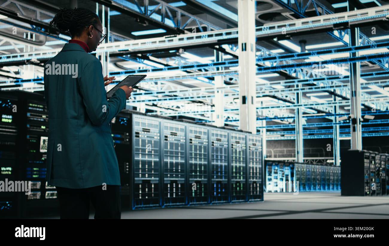 Female programmer walking in data center using tablet to write code, compiling software patches. African american woman in server hub using device application to run programming script, camera A Stock Photo