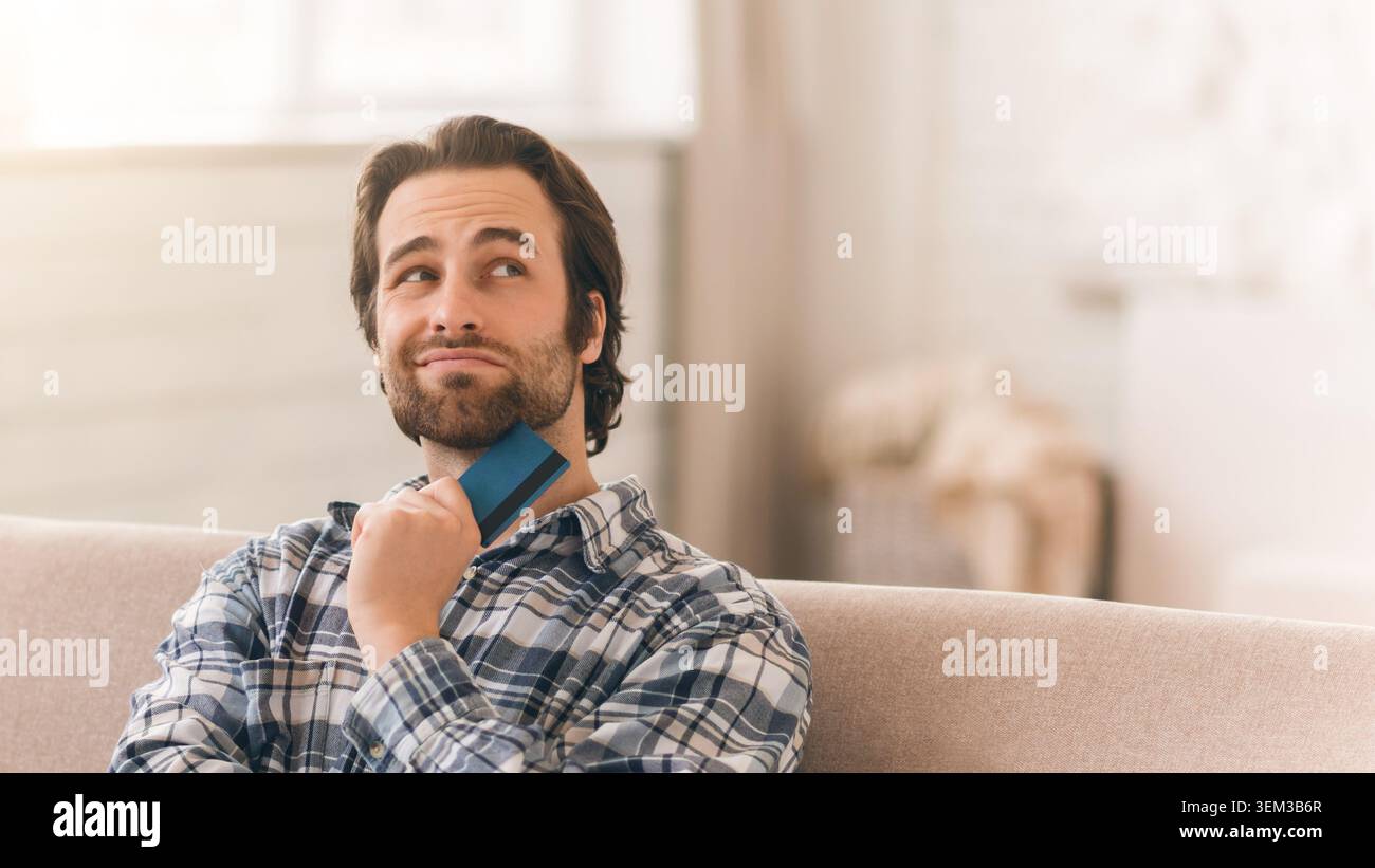 Thoughtful Caucasian Man Holding Credit Card, Considering Online Shopping At Home, Business Concept Stock Photo