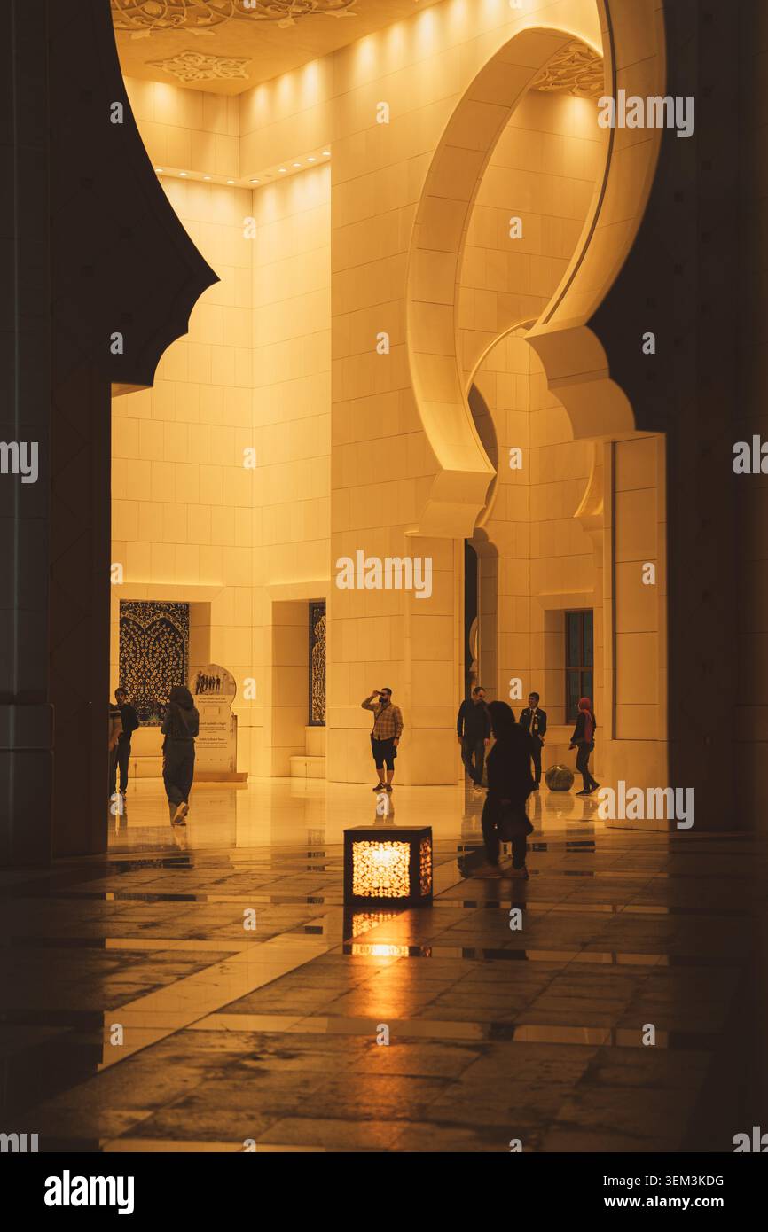 Warmly lit mosque interior with tall arches, reflective floor and people walking through the grand hall. Stock Photo