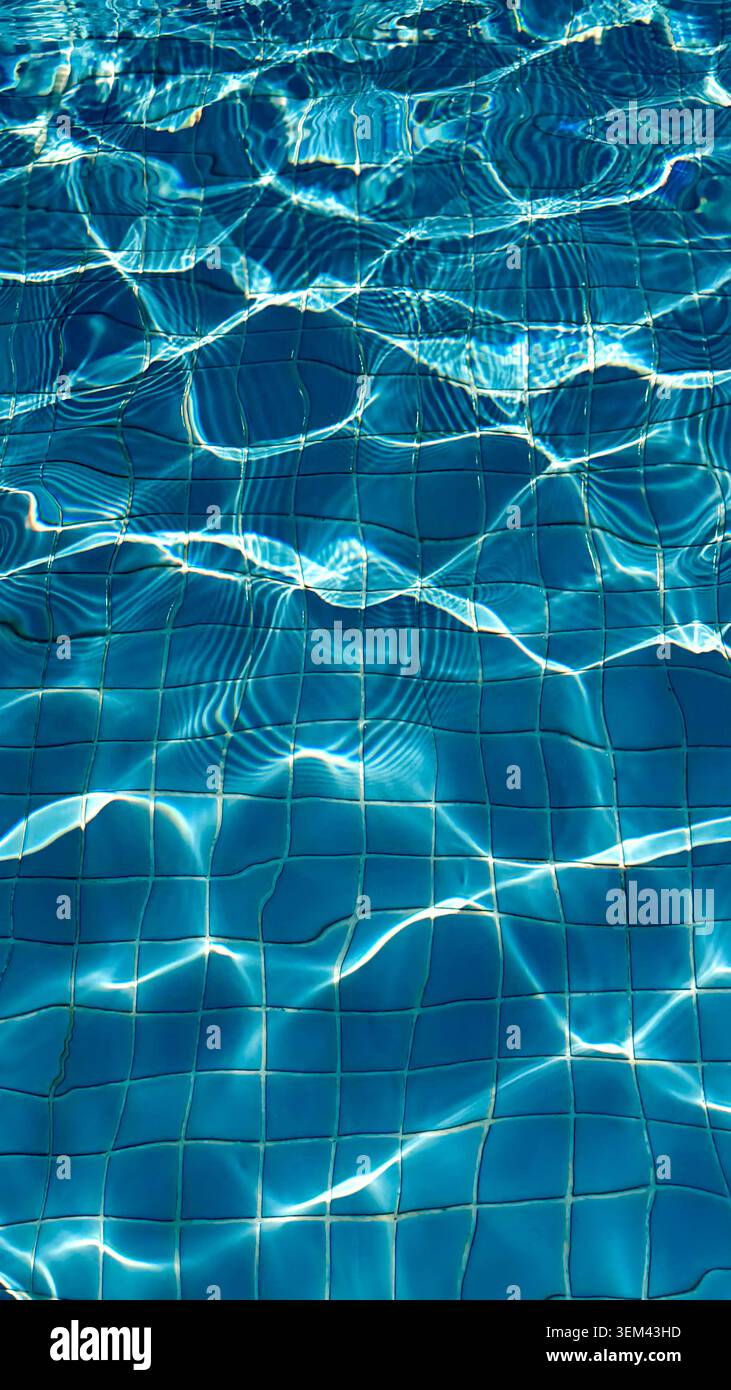 Clear blue swimming pool water surface with sunlight reflections creating ripples and patterns over white tiled floor beneath Stock Photo