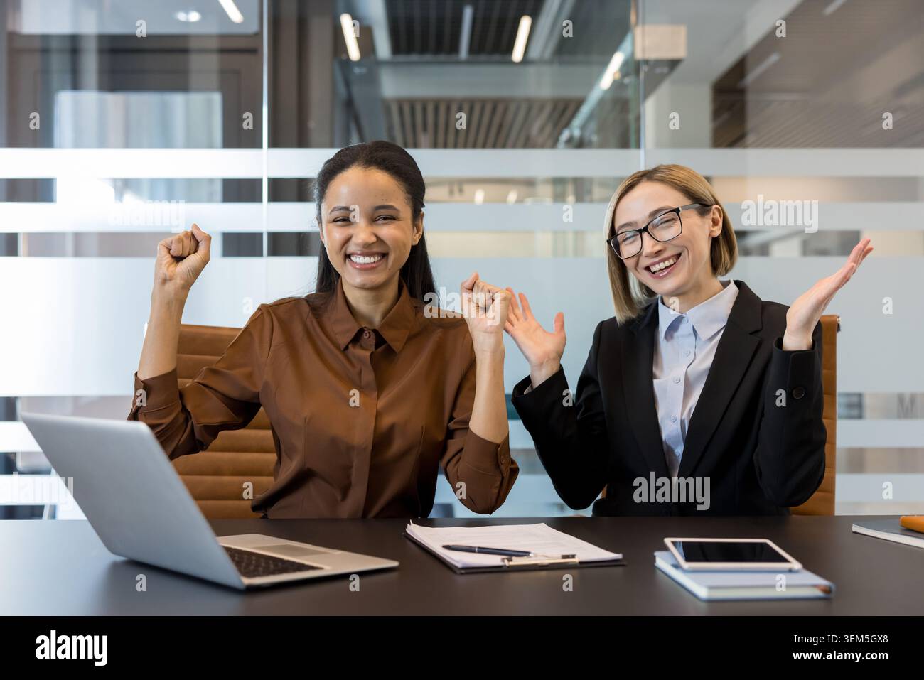 Two happy diverse business women celebrating their success, looking at the camera with arms raised and wide smiles while working together in a modern office environment Stock Photo