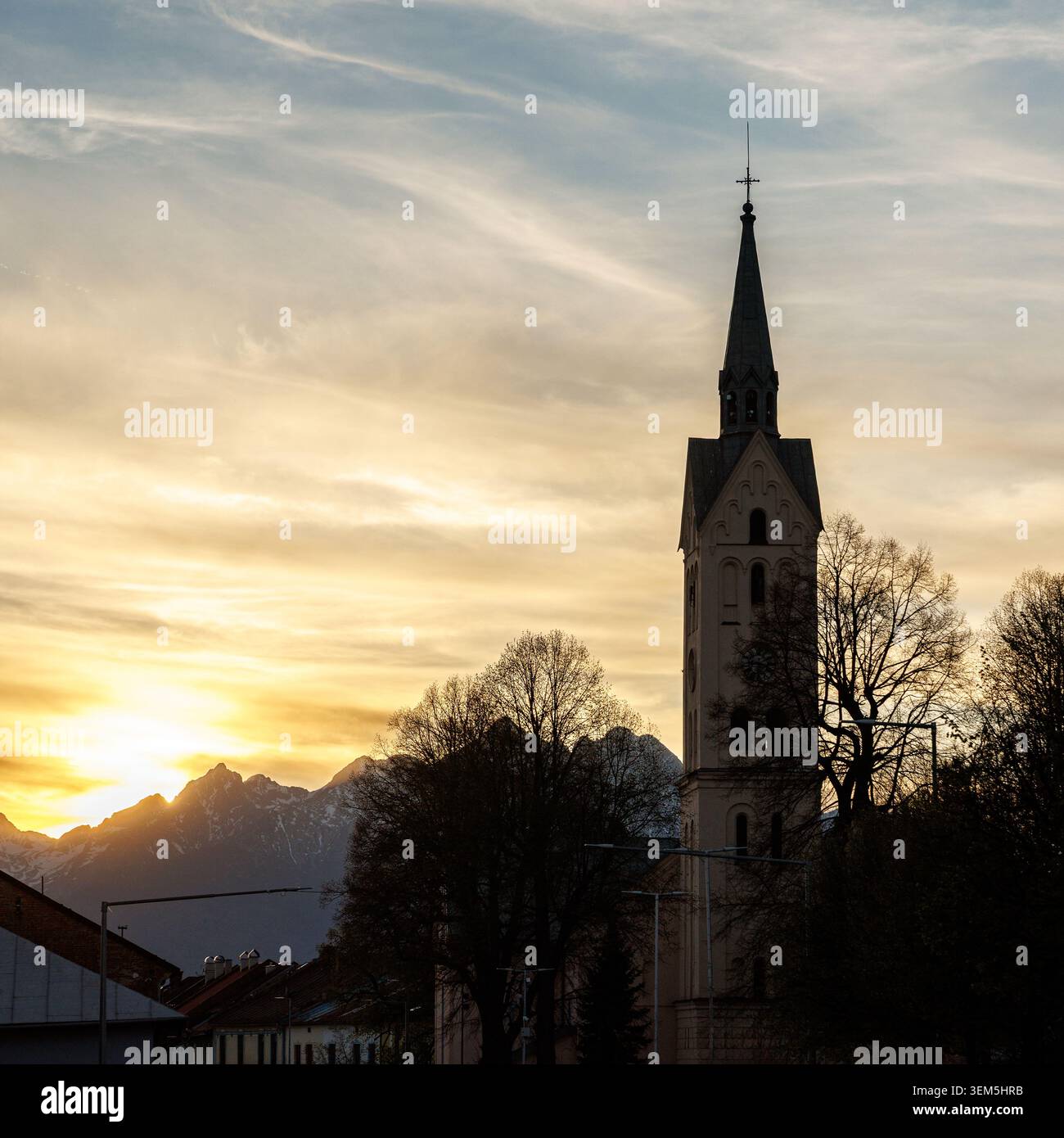An elegant cathedral spire rises prominently in the foreground with the sharp, snow-dusted peaks of a mountain range glowing under a golden dusk sky. Stock Photo