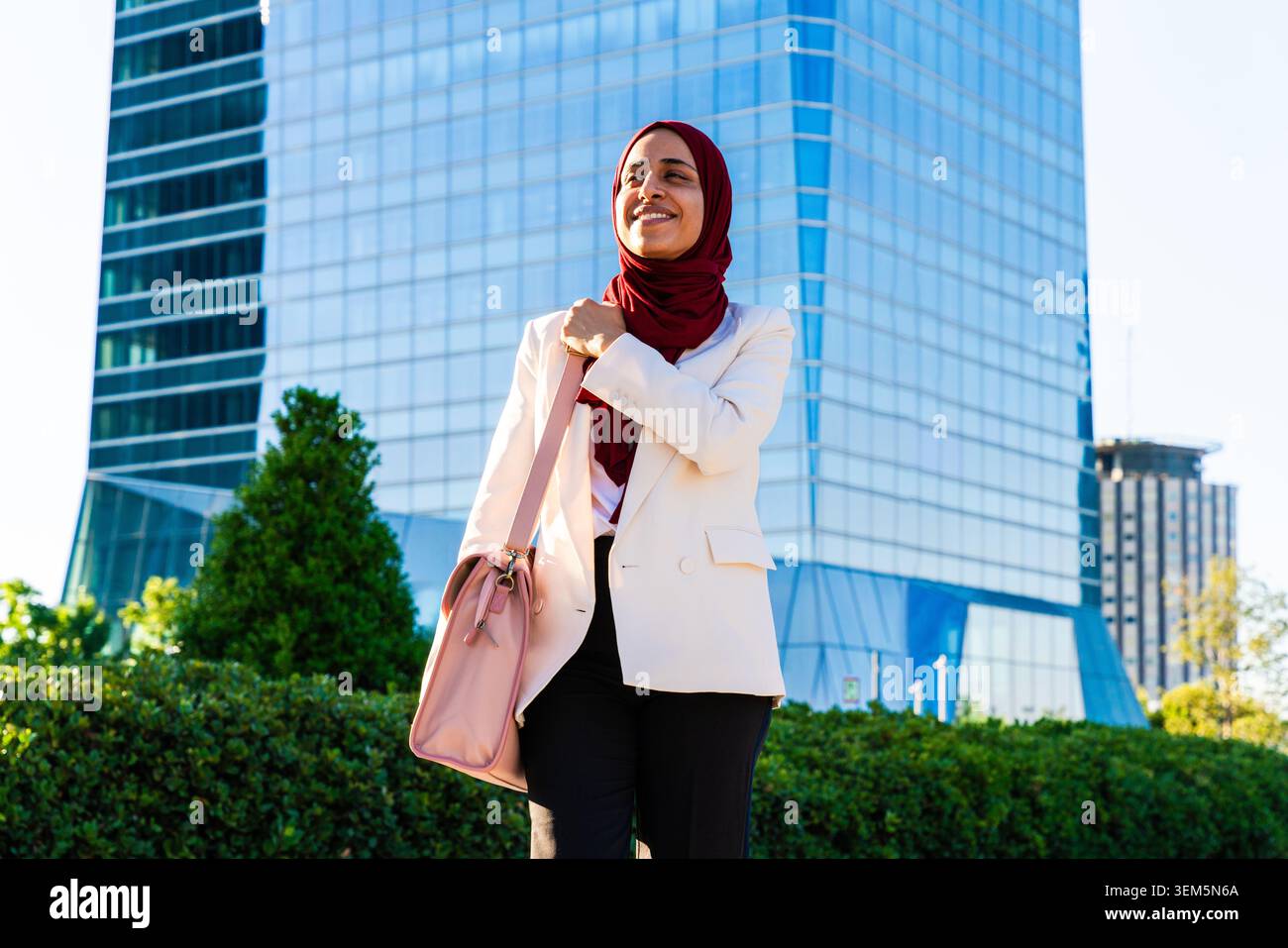 Middle-eastern arab businesswoman wearing hijab strolling in the city - young woman smiling, confidently carrying a bag, walking towards a corporate b Stock Photo
