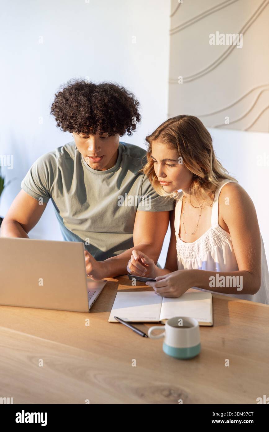 Couple wearing muted green tee and white top, sitting in home workspace using laptop and smartphone Stock Photo
