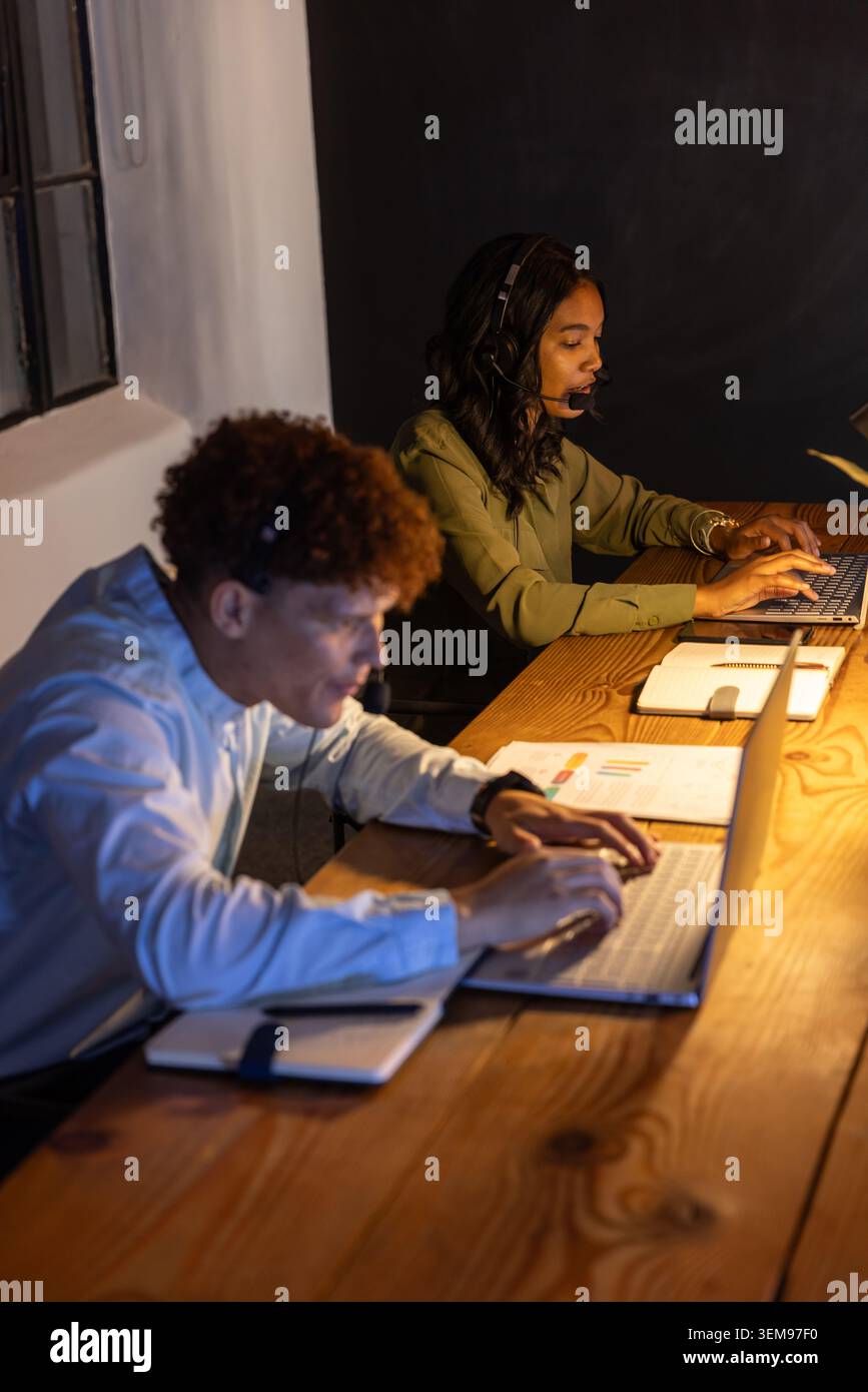 Diverse coworkers working at wooden desk in office wearing headsets and typing on laptops Stock Photo