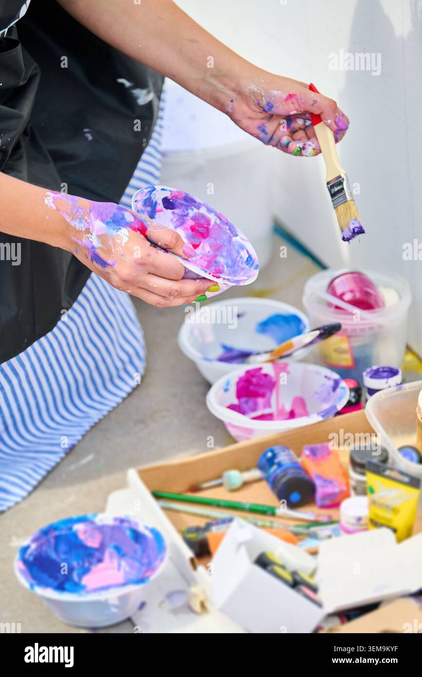 Artists hands covered in mixed paint holding brush and palette bowl with vibrant pink and purple hues, surrounded by various mixing bowls filled with Stock Photo