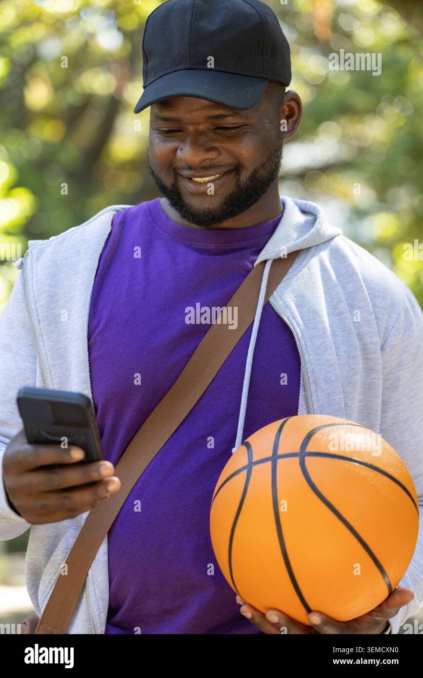 African American man holding orange basketball and checking smartphone in sunny park with bag strap Stock Photo