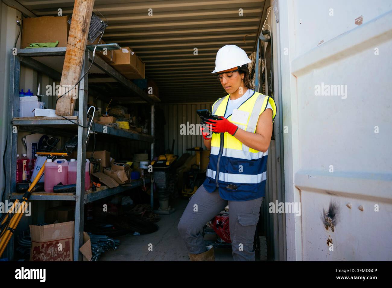 A female construction worker in hard hat and reflective vest stands in a storage container, checking a smartphone among tools and supplies at an activ Stock Photo