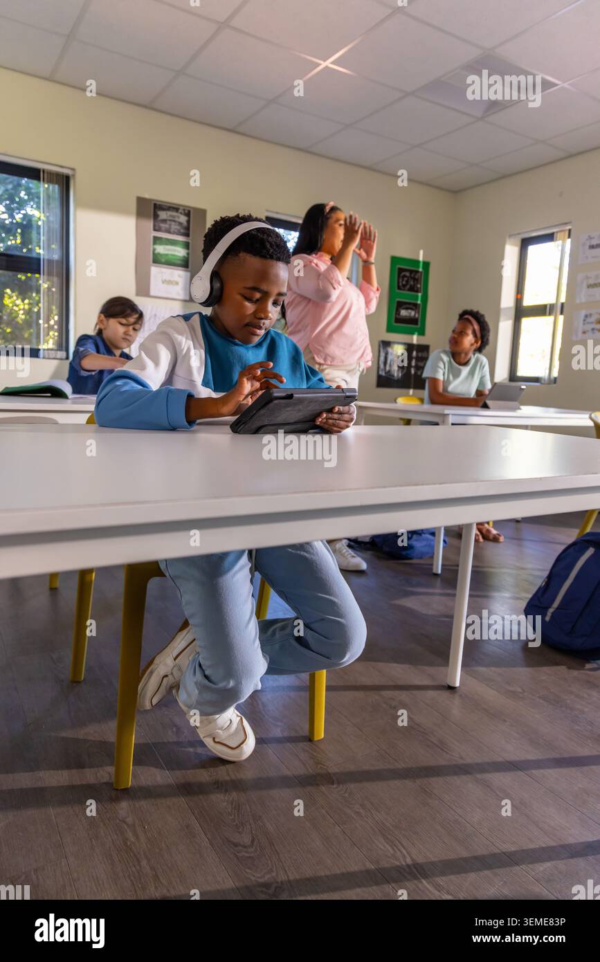 Diverse students with female teacher gesturing in classroom while boy wearing headset using tablet Stock Photo
