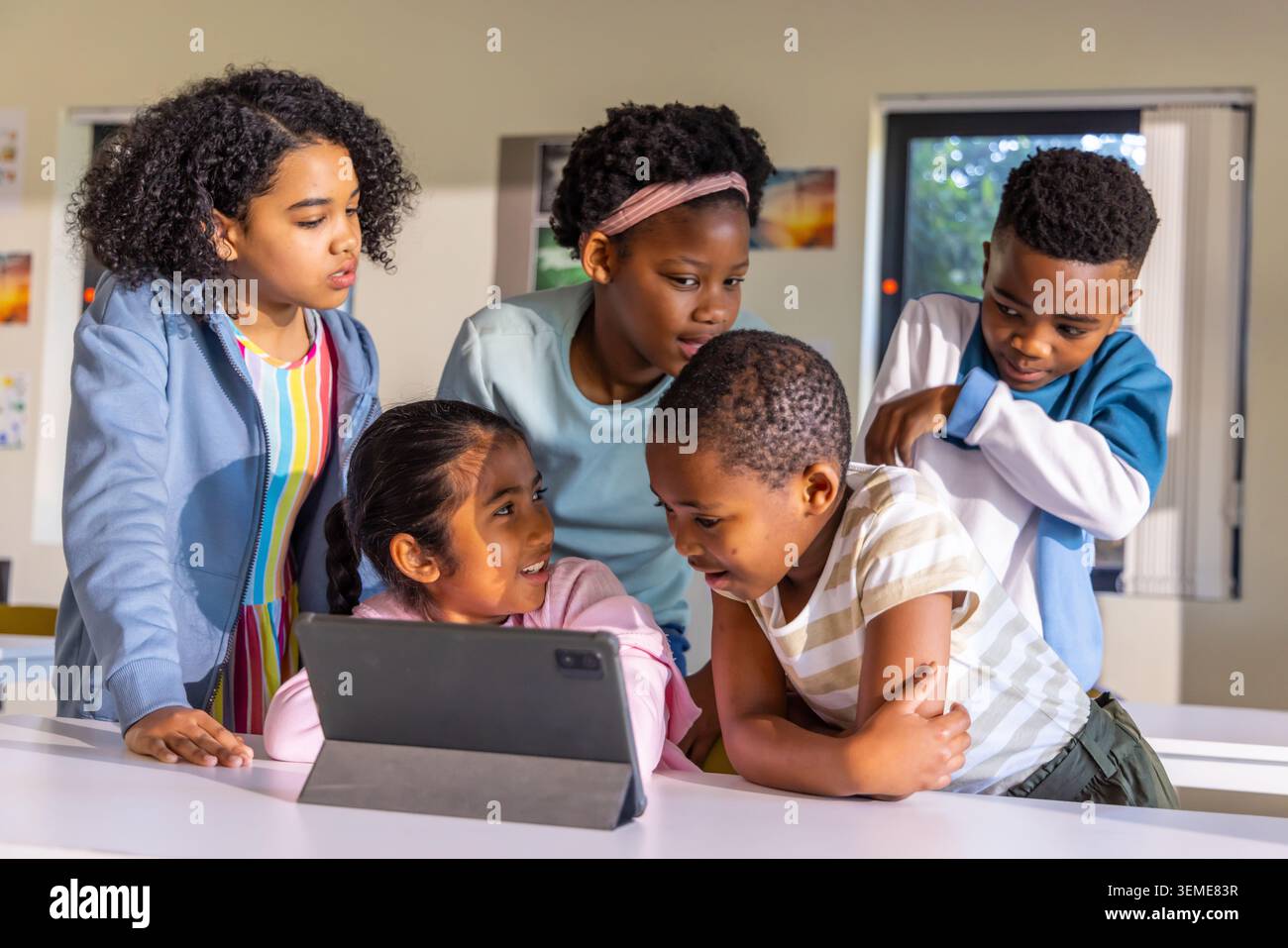 Diverse children gathering around tablet in folio on white classroom table, leaning forward Stock Photo