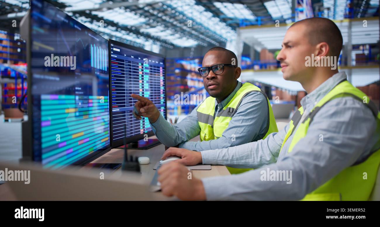 Two Workers Sitting In Warehouse Using Computer For Logistics Management Tasks. Stock Photo