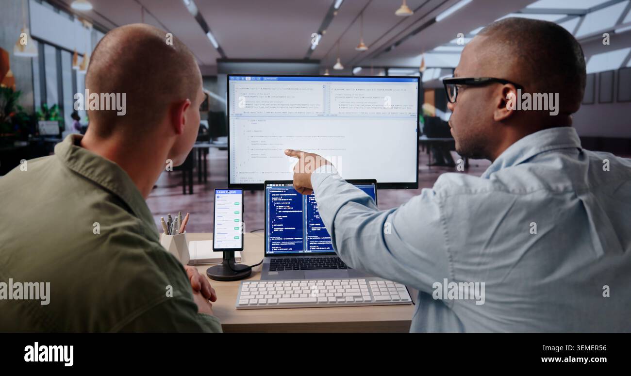 Young Software Engineer Uses Laptop And Desktop To Debug Mobile App. Stock Photo