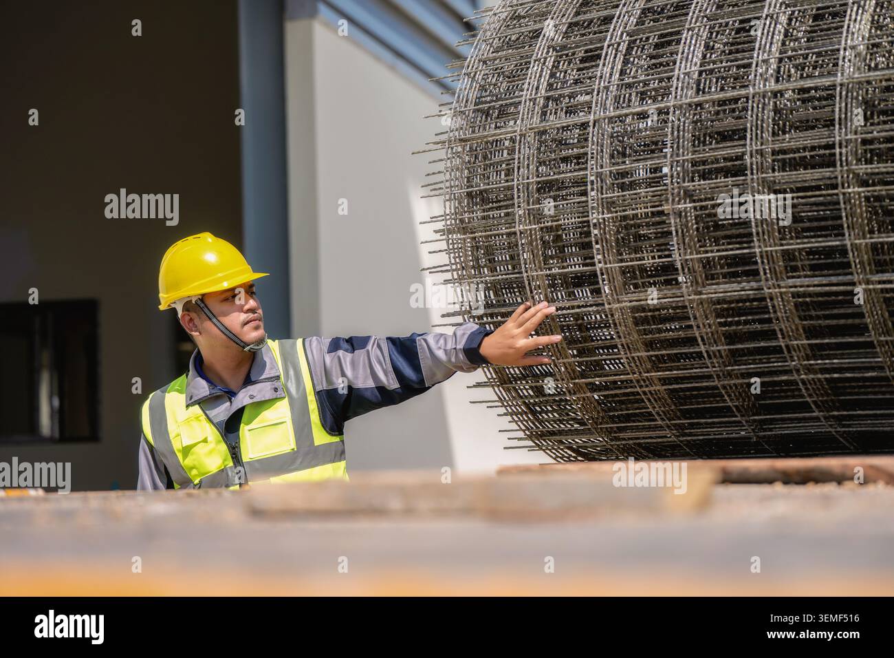 Construction worker pushes large steel mesh roll. He wears yellow hard hat and high-visibility safety vest. Building with windows and concrete backgro Stock Photo