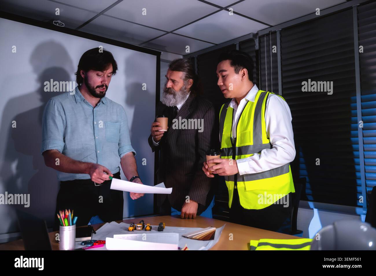 Three people review blueprints late at night, discussing plans. They work in team meeting setting, engineering blueprint review with senior business c Stock Photo
