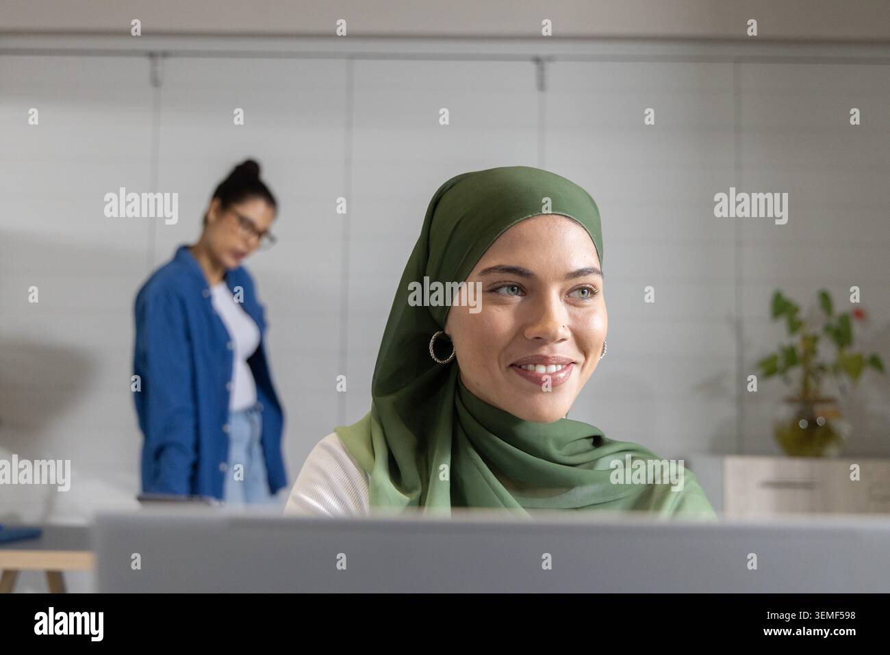 Diverse women team working at desk in office, hijab-wearing woman on laptop, colleague with glasses Stock Photo