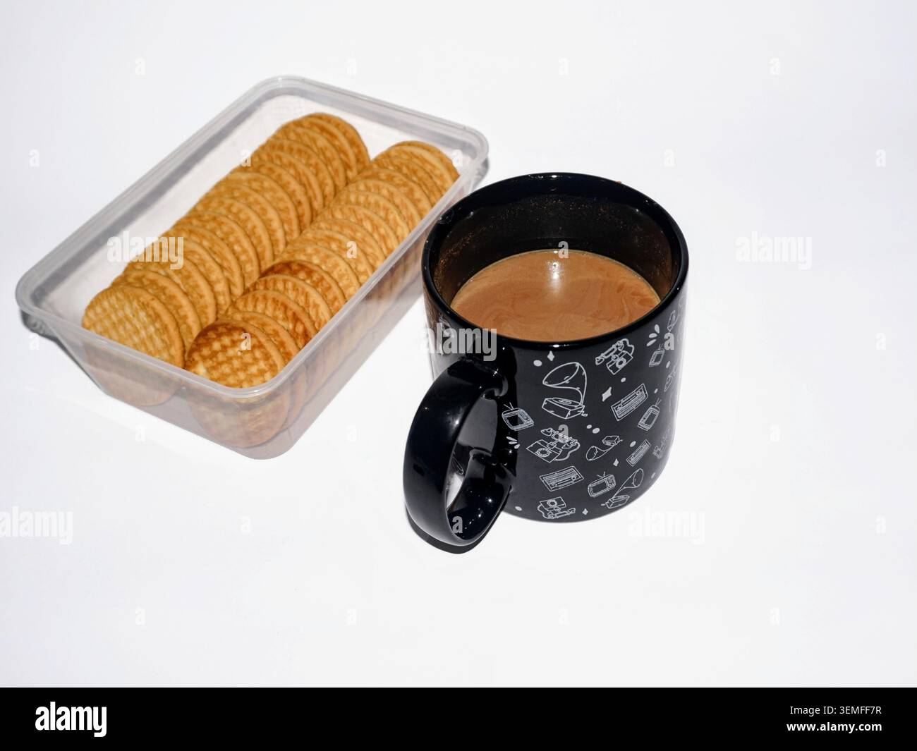 A cup of hot brown milk coffee in a black cup with white retro illustrations, placed next to a clear plastic container containing round biscuits on a bright white background Stock Photo