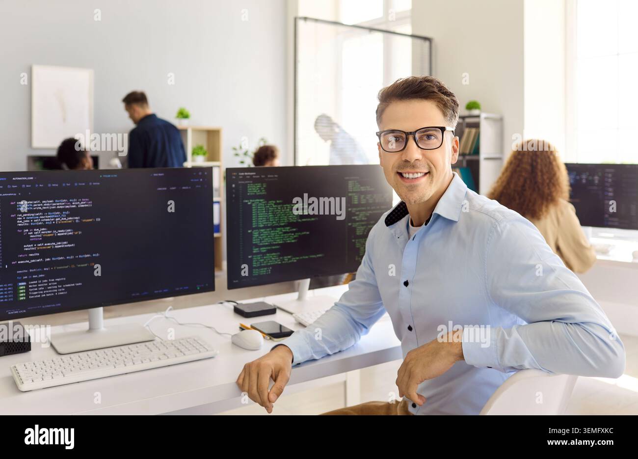 Confident programmer posing by code monitors at desk in bright modern office Stock Photo
