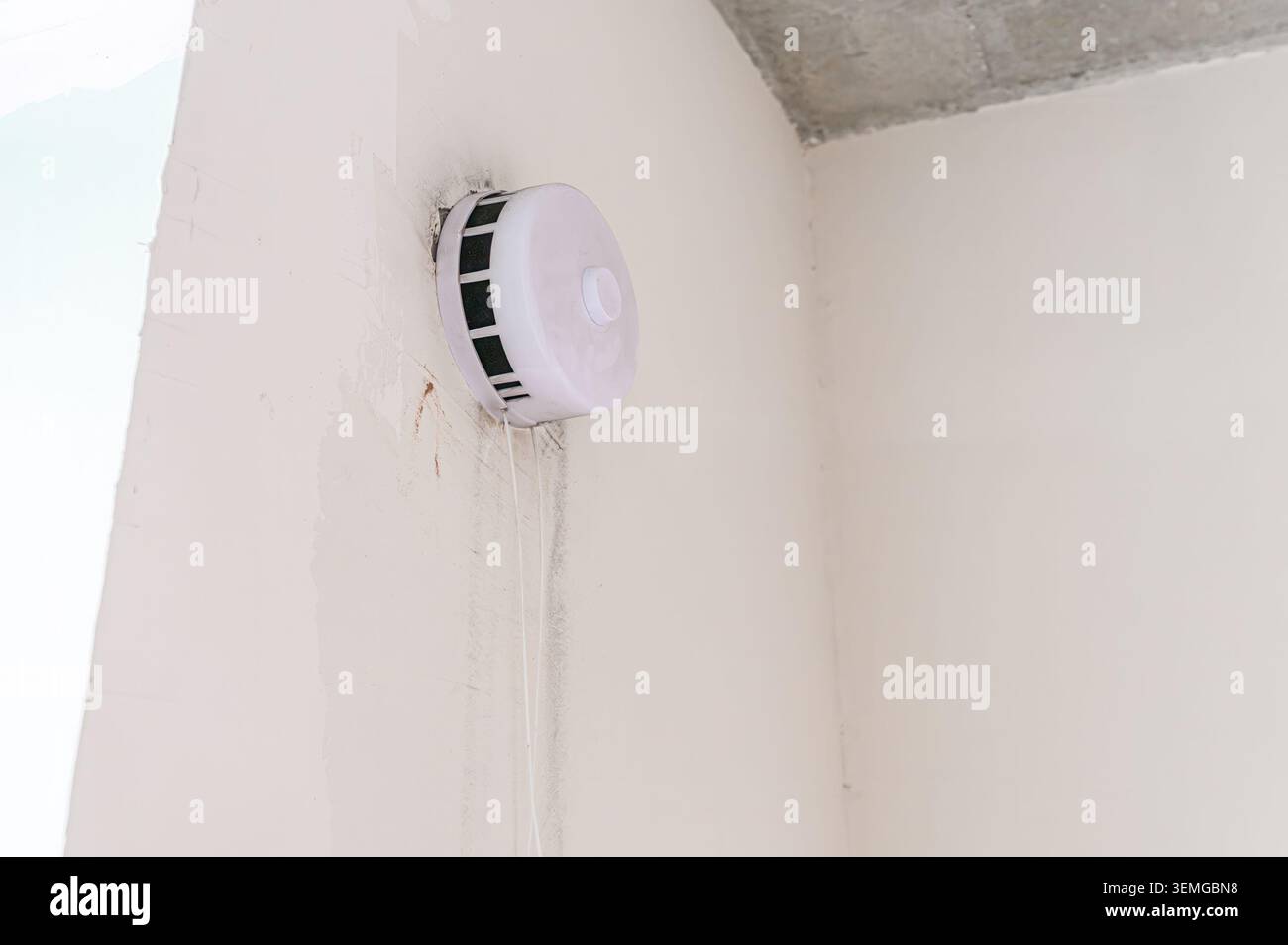 smoke detector is mounted on a white wall in a corner. The wall has some stains and the ceiling is visible Stock Photo