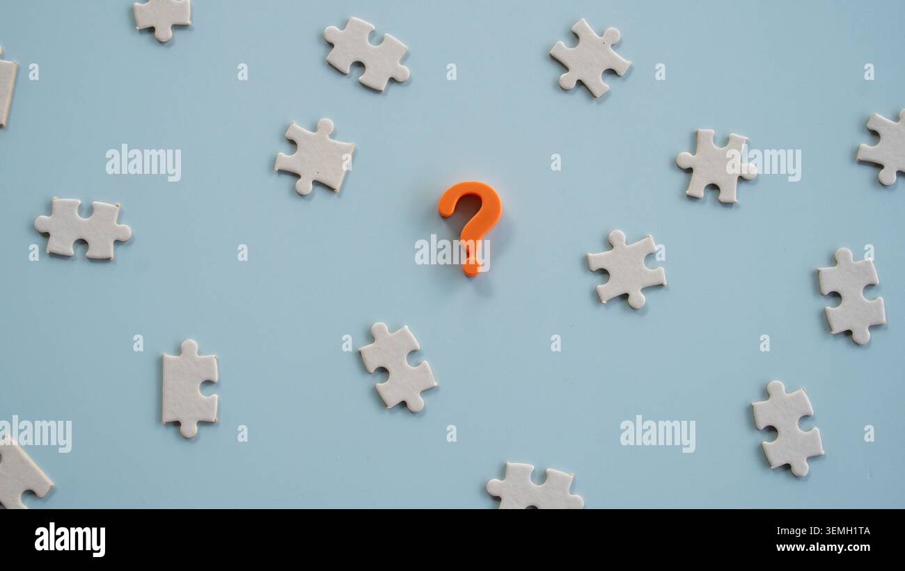 Top view of an orange question mark icon centered among scattered white jigsaw puzzle pieces on a blue background Stock Photo