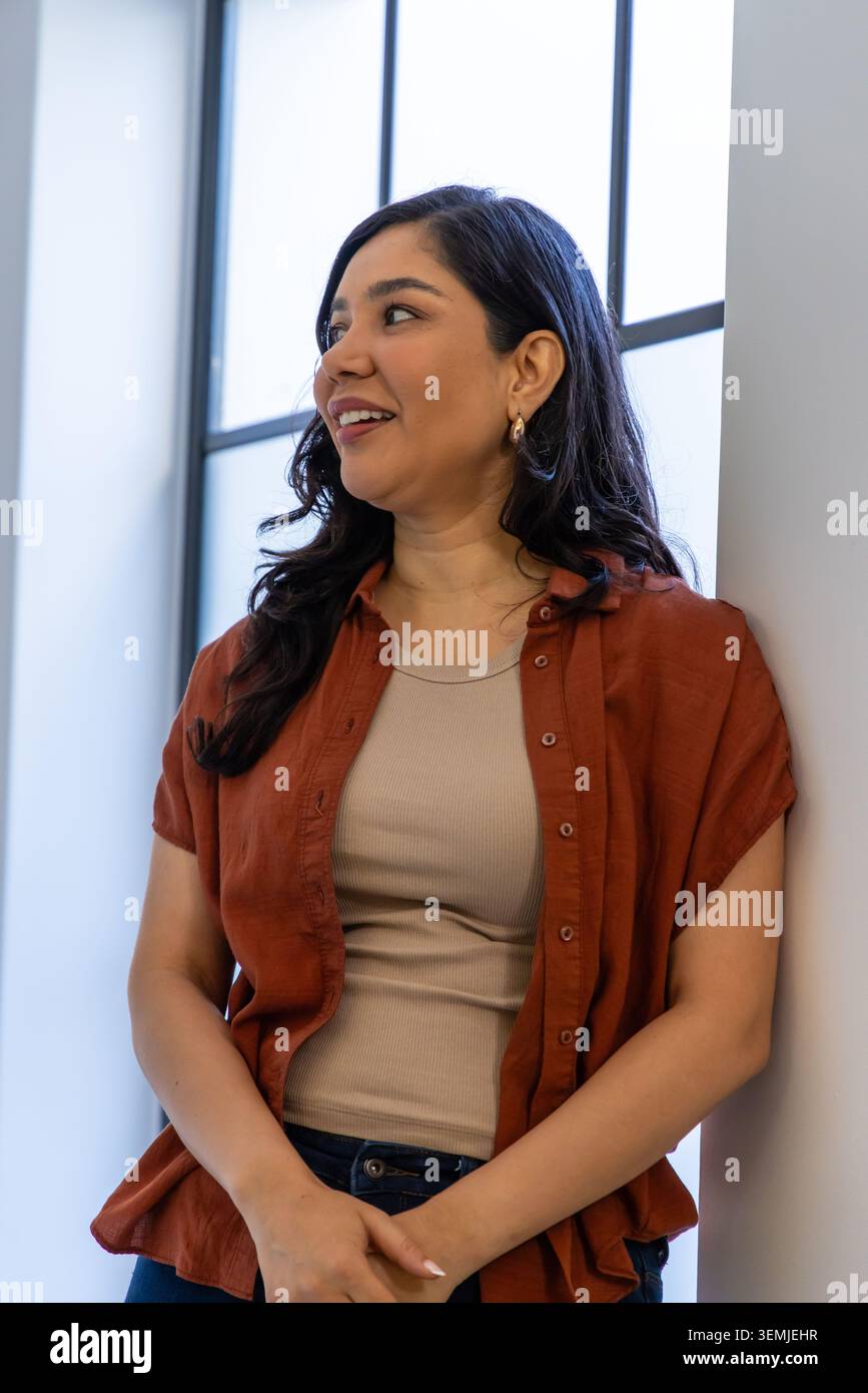 Woman leaning against white wall in studio by tall window, wearing rust shirt and gold hoops Stock Photo