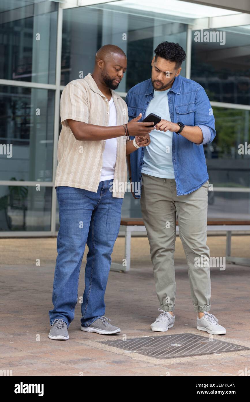 Two African American men standing on paved plaza outside modern glass building checking smartphone Stock Photo