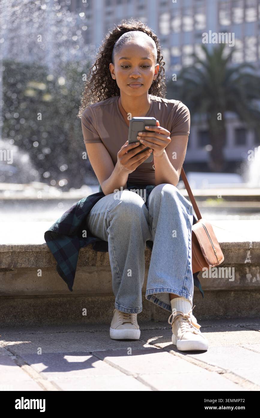 African American woman in her 20s sitting on stone fountain edge holding smartphone, wearing jeans Stock Photo