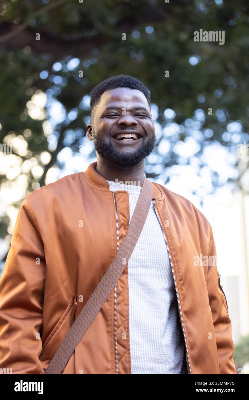 Adult man standing smiling in park, wearing rust bomber jacket, white textured sweater, tan strap Stock Photo