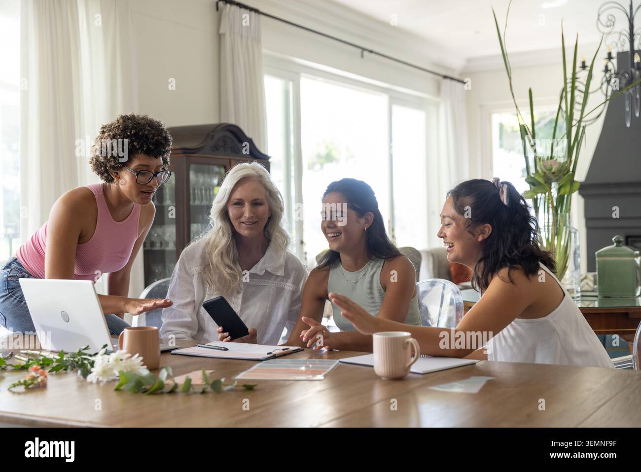 Intergenerational female group reviewing documents, using phone, laptop, tablet at dining table Stock Photo