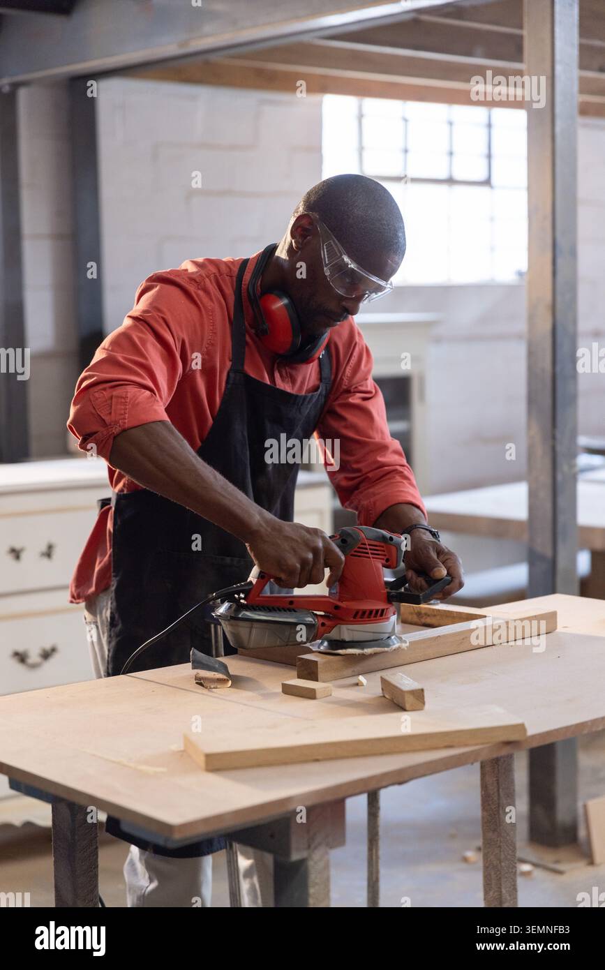 Red electric sander smoothing wooden plank on workbench in woodshop with offcuts Stock Photo