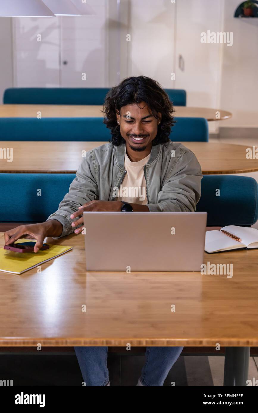 Indian man mid-20s smiling while working on silver laptop in shared study lounge with smartphone Stock Photo